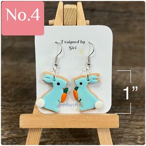 May include: A pair of blue bunny-shaped earrings with orange carrot accents. The earrings are 1 inch in size and are designed by Siri.