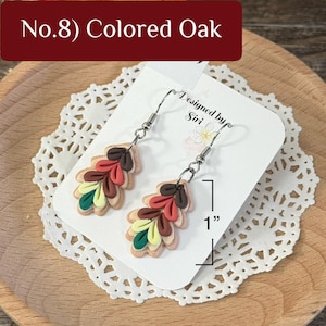 May include: A pair of dangle earrings featuring layered, leaf-shaped charms in dark brown, red, brown, yellow, and green. The earrings are attached to a white display card labeled No.8 Colored Oak and Designed by Siri, showing a 1 inch measurement.