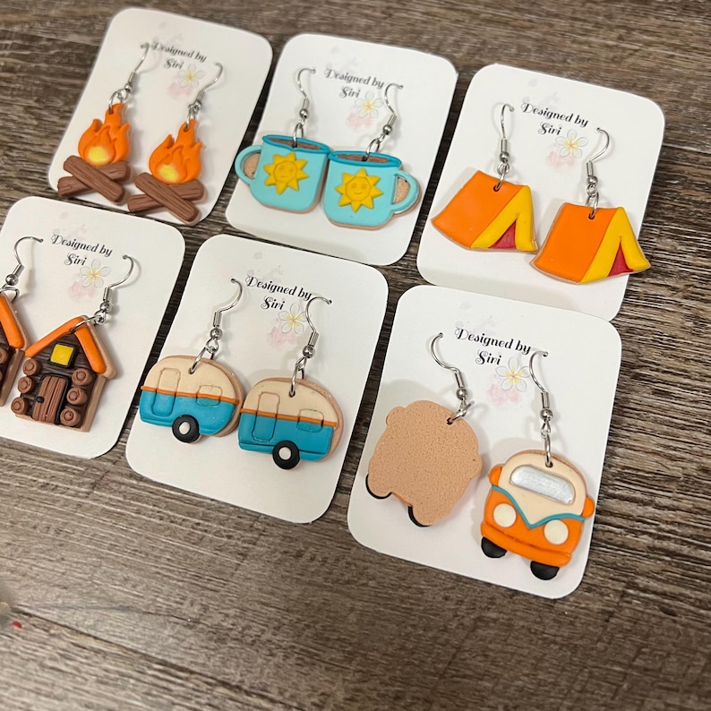 May include: A collection of handmade earrings with camping themes. Designs include campfires, mugs with suns, orange tents, a cabin, blue and tan campers, and an orange van. Each earring set is displayed on a white card with the text "Designed by Siri".