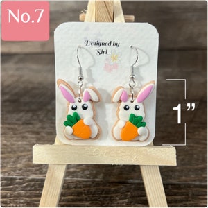 May include: A pair of white bunny rabbit dangle earrings with pink ears and holding a carrot. The earrings are 1 inch long and are designed by Siri.