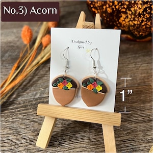 May include: A pair of acorn-shaped dangle earrings featuring a dark brown cap adorned with small orange, yellow, and pink flowers and green leaves, attached to silver-tone hooks. The earrings measure 1 inch in height on a display card.
