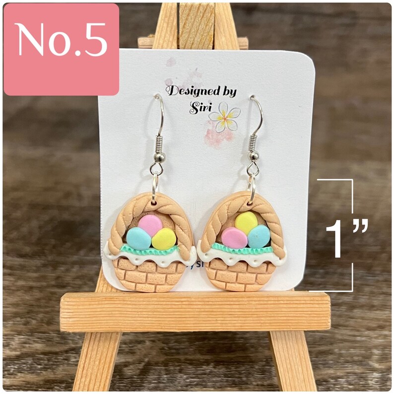 May include: A pair of dangle earrings featuring a brown basket with pastel yellow, blue, and pink Easter eggs. The earrings are 1 inch long. Designed by Siri.