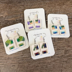 Puede incluir: Pendientes hechos a mano de arcilla polimérica con estanterías en miniatura con libros y plantas. Cada pendiente tiene un marco blanco y un gancho plateado. Diseñado por Siri.