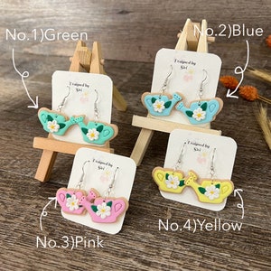 May include: Four pairs of polymer clay earrings shaped like watering cans with flowers. The earrings are displayed on small easels. The colors of the earrings are green, blue, pink, and yellow. The text "Designed by Siri" is on each card.