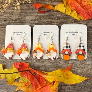 May include: Three pairs of fall-themed gnome earrings. Each gnome has a white beard, a colorful hat, and a unique design, including leaves, acorns, and pumpkins. The earrings are displayed on white cards with the text "Designed by Siri".
