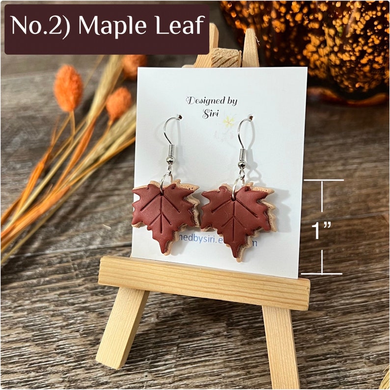 May include: A pair of maple leaf earrings featuring dark brown leaf-shaped charms with etched veins, suspended from silver-toned metal hooks. The earrings are displayed on a white card labeled No.2 Maple Leaf, showing a 1 inch height.