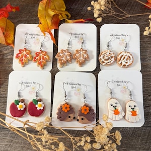 May include: Six pairs of dangle earrings on white display cards, featuring autumn-themed polymer clay charms: orange maple leaves, brown gingerbread leaves, cinnamon rolls, red apples with daisies, brown hedgehogs, and white ghosts holding pumpkins.