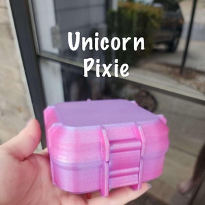 May include: A small, square, pink and purple box with a textured surface. The box has the words "Unicorn Pixie" written on it.