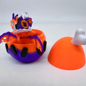 May include: A 3D-printed toy set featuring a dragon-like creature emerging from an orange and purple container. The creature has orange, purple, and white accents. A separate orange dome with a white bone-shaped handle is also present.