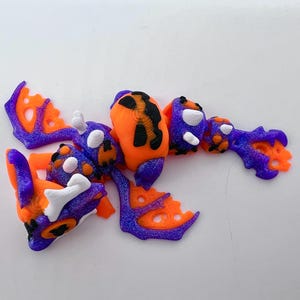 May include: A collection of colorful, 3D-printed dragon or monster parts in orange, purple, and white. The pieces include wings, body segments, and head components, all with intricate details and a textured appearance.