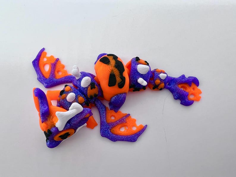 May include: A collection of colorful, 3D-printed dragon or monster parts in orange, purple, and white. The pieces include wings, body segments, and head components, all with intricate details and a textured appearance.