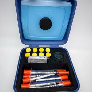 May include: A blue plastic case with a thermometer reading 88 degrees Fahrenheit. The case contains a blue ice pack, vials with yellow tops, sterile alcohol prep pads, and several syringes with orange tips.