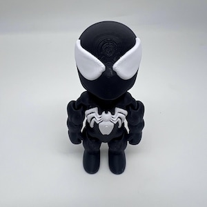 May include: A black and white figurine of a superhero character. The figure has a round head with white eye shapes and a white spider emblem on the chest. The figure is standing on a white surface.