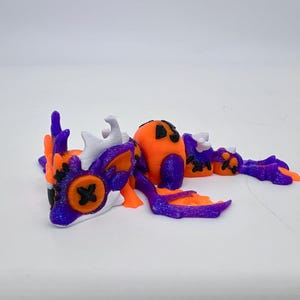 May include: A 3D-printed dragon figurine in orange, purple, and white. The dragon has a pumpkin-shaped head and body, with black cross-shaped eyes. The wings and tail are purple with orange accents. The figurine is approximately 6 inches long.