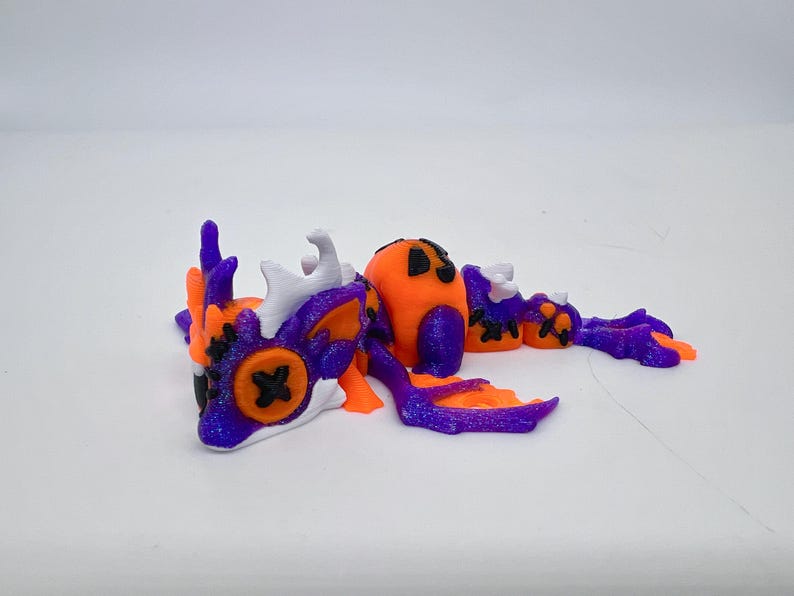 May include: A 3D-printed dragon figurine in orange, purple, and white. The dragon has a pumpkin-shaped head and body, with black cross-shaped eyes. The wings and tail are purple with orange accents. The figurine is approximately 6 inches long.