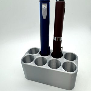 May include: A silver 3D printed pen holder with six slots, two of which are filled with blue and brown pens.