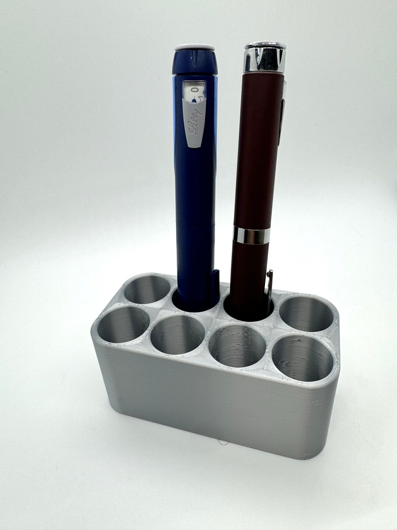 May include: A silver 3D printed pen holder with six slots, two of which are filled with blue and brown pens.
