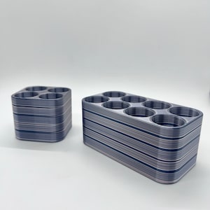 May include: Two 3D printed storage organizers with a striped pattern in shades of gray and white. The organizers have multiple circular openings for holding small items.