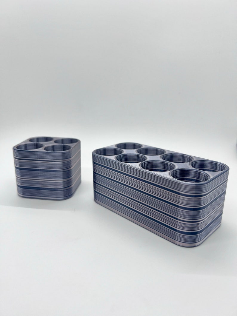 May include: Two 3D printed storage organizers with a striped pattern in shades of gray and white. The organizers have multiple circular openings for holding small items.