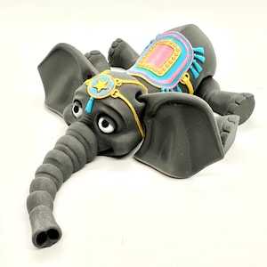 May include: A gray elephant figurine with large ears, a long trunk, and big eyes. The elephant has a colorful saddle with pink, blue, and yellow accents. A gold-colored star and blue tassel adorn the elephant's head.