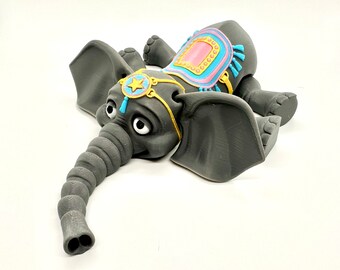 May include: A gray elephant figurine with large ears, a long trunk, and big eyes. The elephant has a colorful saddle with pink, blue, and yellow accents. A gold-colored star and blue tassel adorn the elephant's head.
