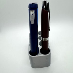 May include: A gray plastic pen holder with six slots, holding a blue and white insulin pen and a brown pen.