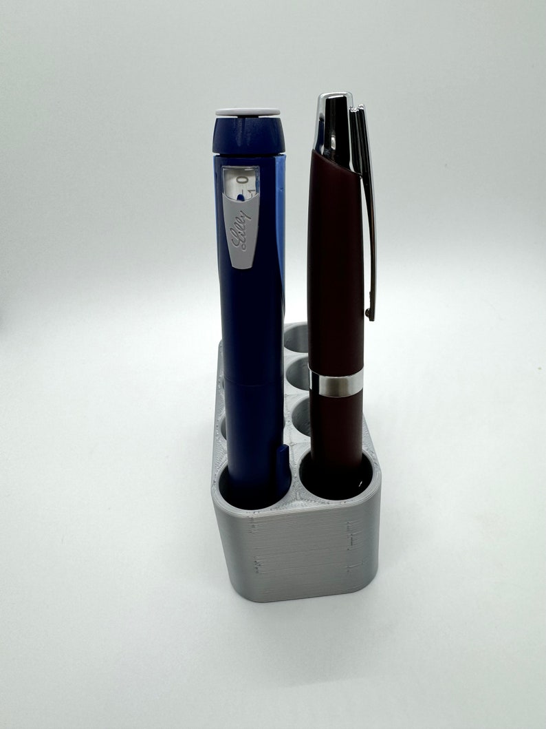 May include: A gray plastic pen holder with six slots, holding a blue and white insulin pen and a brown pen.