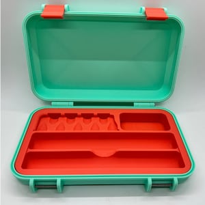 May include: A mint-green rectangular case, open to reveal a bright orange interior with compartments. The case has a hinged lid and orange clasps. The interior includes slots and storage areas. The case is designed for carrying small items.