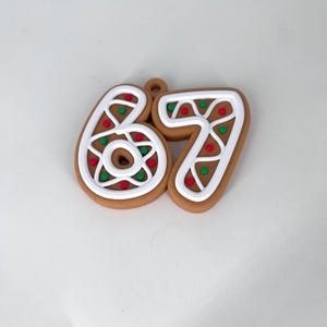 May include: A gingerbread-style ornament in the shape of the numbers 67. The numbers are brown with white icing and decorated with red and green sprinkles. The ornament has a small loop at the top for hanging.