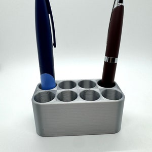 May include: A silver pen holder with eight slots, two pens are in the holder, one is blue and the other is brown.