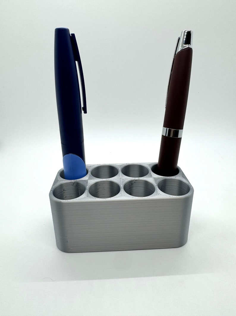 May include: A silver pen holder with eight slots, two pens are in the holder, one is blue and the other is brown.