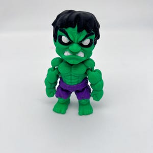 May include: A green Hulk figurine with black hair, white eyes, and a stern expression. The figure is wearing purple shorts. The toy is standing on a white surface.