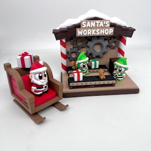 May include: A Christmas-themed diorama featuring Santa Claus in a sleigh and a workshop scene. The workshop has the text "SANTA'S WORKSHOP" on the roof. Elves are working on a conveyor belt with gifts. The sleigh has a red seat and is loaded with gifts.