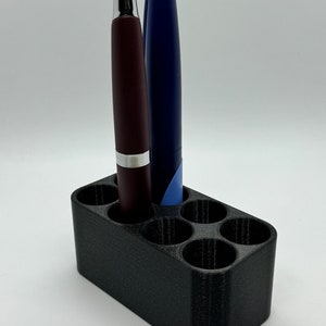 May include: A black plastic pen holder with six slots, holding two pens, one burgundy and one blue.