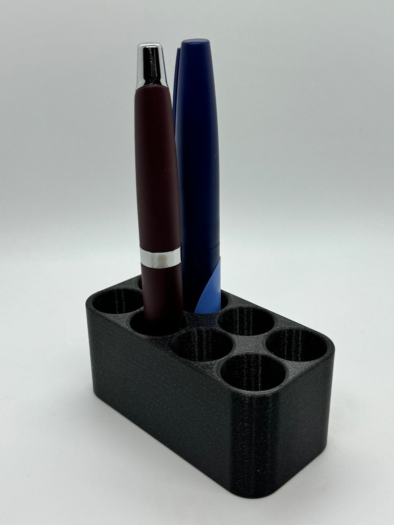 May include: A black plastic pen holder with six slots, holding two pens, one burgundy and one blue.