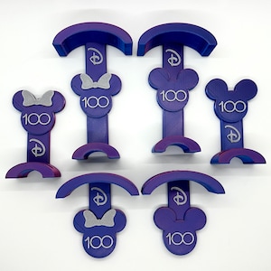 May include: Six purple and silver Mickey Mouse and Minnie Mouse themed 100th anniversary Disney character display stands. Each stand features a Mickey or Minnie Mouse head with a bow and the number 100. The stands are designed to hold small items, such as pins or charms.