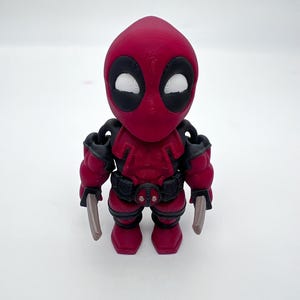 May include: A 3D-printed Deadpool figurine. The figure is primarily red with black accents, including the eyes, belt, and shoulder armor. It has two silver-colored swords. The figurine is standing upright on a white surface.