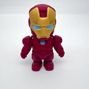 May include: A 3D-printed Iron Man figurine. The figure is primarily red with a gold-colored mask and blue eye accents. The figurine has a stylized, blocky design, with visible layer lines from the 3D printing process. The figure is standing upright.