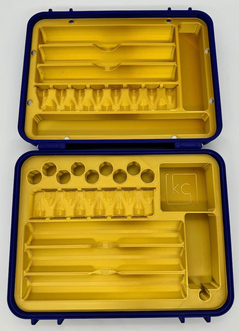 May include: A blue plastic case with a gold interior. The case has multiple compartments for storing small items. The case has a label with the letters "KC" on it.