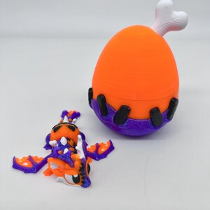 May include: An orange and purple toy dragon with outstretched wings and a matching orange and purple egg-shaped container with a white bone-shaped handle. The dragon has white accents and black eyes. The container has black accents.
