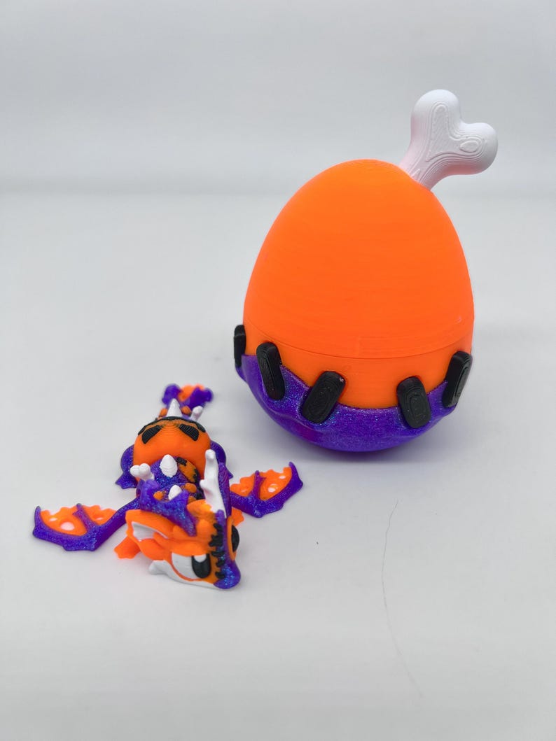 May include: An orange and purple toy dragon with outstretched wings and a matching orange and purple egg-shaped container with a white bone-shaped handle. The dragon has white accents and black eyes. The container has black accents.