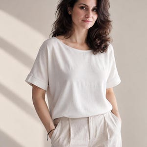May include: A short-sleeved, off-white blouse with a wide, rounded neckline and cuffed sleeves, paired with light beige trousers featuring front pleats. The blouse has a relaxed fit and a smooth, solid-colored fabric texture throughout the garment.
