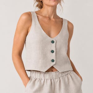 Puede incluir: Un chaleco beige sin mangas con escote en V y tres botones verde oscuro, combinado con pantalones cortos beige a juego con cintura elástica y dobladillos vueltos. El conjunto está confeccionado en un tejido texturizado de aspecto natural.