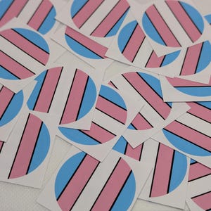May include: A pile of circular stickers displaying the transgender pride flag. Each sticker has a light blue circle with pink and white stripes, and black vertical lines. The stickers are arranged on a white background.