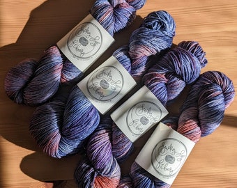 Puede incluir: Cuatro ovillos de hilo teñido a mano en tonos de morado, azul y naranja. El hilo está etiquetado como "Lucky Sky Fibers".