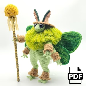 Puede incluir: Criatura de polilla de crochet verde y marrón con un bastón amarillo y marrón. La polilla tiene grandes alas verdes y un cuerpo verde y marrón peludo. El bastón tiene un pompón amarillo en la parte superior. PDF.