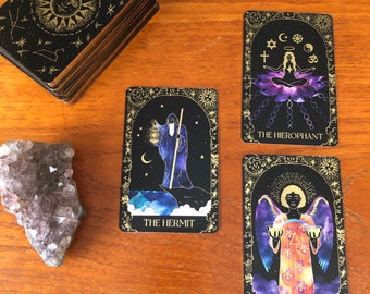 May include: A deck of tarot cards with a black and gold celestial design, featuring the sun, moon, and stars. Three individual cards are displayed, along with a piece of amethyst. The cards depict The Hermit, The Hierophant, and Temperance.