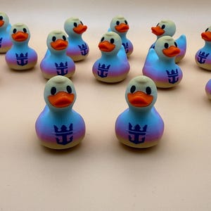 May include: A collection of small rubber ducks featuring a gradient color pattern of pink, yellow, and blue. Each duck has an orange beak and a dark blue anchor and crown logo printed on the chest. The ducks are arranged in a group on a surface.