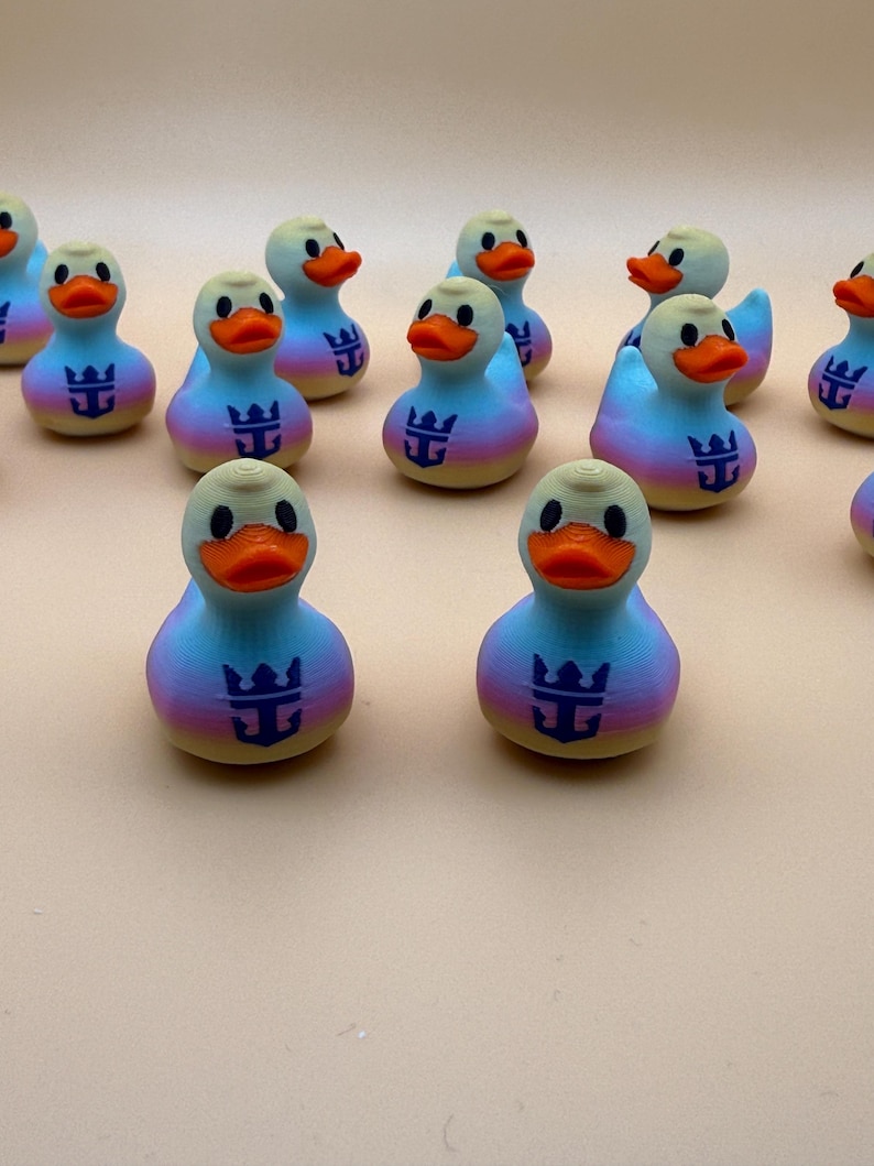 May include: A collection of small rubber ducks featuring a gradient color pattern of pink, yellow, and blue. Each duck has an orange beak and a dark blue anchor and crown logo printed on the chest. The ducks are arranged in a group on a surface.