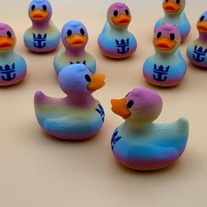 May include: A collection of small, 3D-printed rubber ducks featuring a gradient rainbow color scheme. Each duck has an orange beak, black eyes, and a dark blue anchor-like logo printed on its chest. The ducks are arranged in a group.